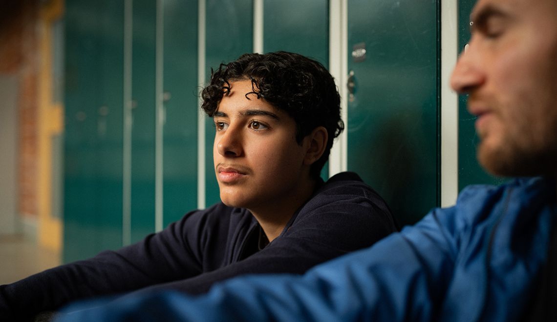 En ung person och en vuxen man sitter och samtalar framför skolskåp i en skola.